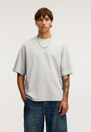 Szürke, oversized póló, puha anyagból készült, rövid ujjú és kerek nyakkivágással. Sötétkék farmernadrággal viselve, az egyik karon tetoválások láthatók.