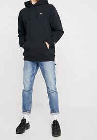 Sudadera negra de algodón con cordones, bolsillo frontal, combinada con vaqueros de mezclilla azul claro y zapatillas deportivas negras. Se ven calcetines blancos hasta el tobillo.