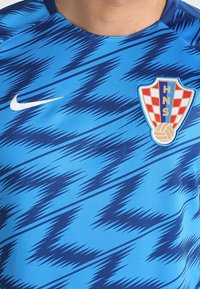 Camiseta atlética azul con un patrón en zigzag en azul más oscuro, con un logo de Nike en blanco y un parche en forma de escudo con un diseño de cuadros rojos y blancos.