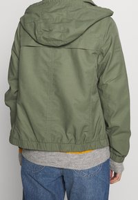 Olivengrøn jakke med hætte, elastisk talje og ribstrikkede manchetter, lagdelt over en grå sweater og blå denimjeans. Enkelt design.