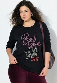 Schwarzes Langarmshirt mit mehrfarbigem Aufdruck "Glaube an dich selbst." Getragen mit burgunderfarbenen Hosen und einer strukturierten Handtasche.
