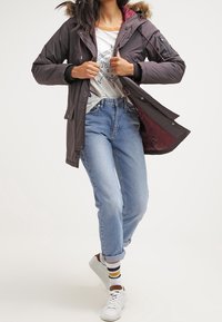 Woman wearing a dark parka with fur hood, white graphic t-shirt, cuffed blue jeans, striped socks, and white sneakers, fastening the jacket.