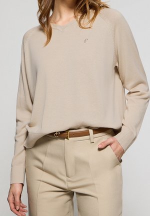 Femme portant un pull beige à col en V et un pantalon beige assorti avec une ceinture marron, une main dans la poche et les cheveux lâchés.