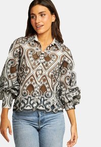 Weiße Bluse mit Muster, die komplexe schwarze und braune Designs zeigt, einen Kragen hat und voluminöse Ärmel mit elastischen Bündchen besitzt. Kombiniert mit blauen Jeans.