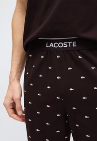 Κοντινό πλάνο ατόμου που φοράει μαύρο παντελόνι Lacoste με λευκά λογότυπα κροκοδείλου και εμφανές λογότυπο στη ζώνη, με το χέρι χαλαρό στο πλάι.