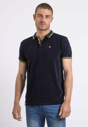Homme portant un polo bleu marine avec des bordures jaunes et un jean bleu clair, debout devant un fond blanc uni.