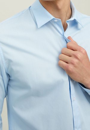 Mand justerer kraven på en lyseblå skjorte med sin højre hånd, så en del af hans hals og øverste torso er synlig.