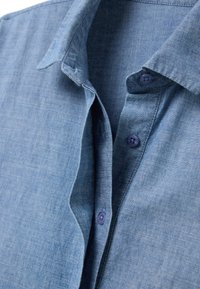 Camicia in chambray azzurro chiaro con una texture morbida, colletto alla francese e bottoni viola. Presenta un design informale e traspirante.