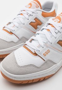 Zapatillas deportivas de cuero blanco con acentos de ante gris, detalles naranjas, secciones perforadas y cordones blancos. Presenta una suela de goma texturizada y la marca "550".