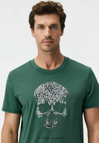 Zielony bawełniany t-shirt z fakturowanym srebrnym motywem czaszki na środku, poniżej tekst "Diverse MCMXCIII". Krótkie rękawy, okrągły dekolt.