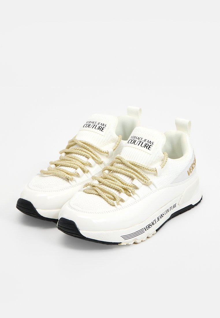 Zapatillas blancas de Versace Jeans Couture con cordones con un patrón dorado, detalles de malla y charol, y texto de marca en la lengüeta, el lateral y la suela.