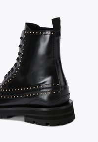 Stivaletto in pelle nera con borchie dorate lungo le cuciture e una suola testurizzata. Presenta lacci e un piccolo passante sul retro per una facile calzata.