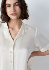 Młoda kobieta z krótkimi brązowymi włosami, nosząca jasnoszeregową bluzkę zapinaną na guziki z krótkim rękawem, stojąca z jedną ręką na biodrze.