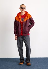 Chaqueta con capucha en color marrón y naranja, camiseta roja, jeans oscuros, zapatos naranjas y negros; el modelo lleva gafas de sol. Tejido suave, ajuste casual.