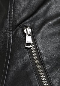 Detalj av en svart skinnjakke med glidelås med metallpull, teksturert overflate og fremhevet søm langs sømmen.