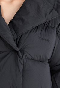Chaqueta de plumas negra con un amplio cuello doblado y detalles de costura. Presenta un sutil logo de Calvin Klein en la tela.
