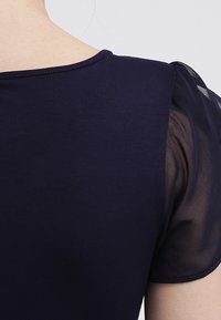 Marinblus med en solid tygkropp och semi-genomskinliga puffärmar. Den släta texturen framhäver halslinningens och ärmsömmarnas detaljer.