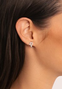 Orecchino a bottone d'argento a forma di stella, con gemme trasparenti incastonate lungo il bordo, indossato sull'orecchio sinistro. Sfondo con tonalità di pelle liscia.