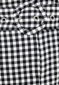 Černobílá kostkovaná látka s kulatým poutkem na pásek, které má stříbrné kovové prvky a kontrastní vertikální a horizontální vzory.