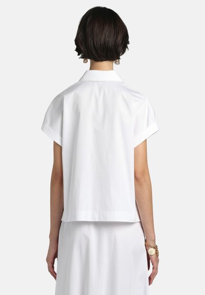 Fehér, rövid ujjú, galléros blúz, dobozszerű szabással és sima textúrával. A hátsó rész enyhén hosszabb, zsebek nélkül.