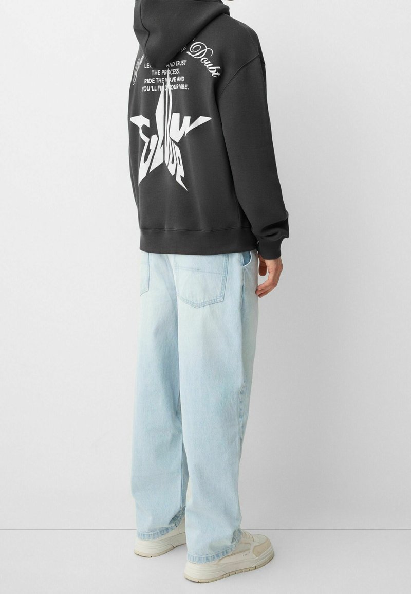 Personne portant un sweat à capuche noir avec une étoile blanche et un texte dans le dos, un jean large bleu clair et des baskets blanches, debout de dos.