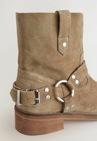 Stivale alla caviglia in suede marrone con fibbia argentata e accentatura circolare, dotato di tacco impilato e cuciture visibili su una superficie testurizzata.