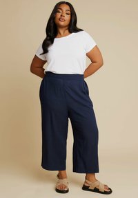 Biała koszulka z krótkim rękawem, granatowe szerokie spodnie z bocznymi kieszeniami oraz beżowe sandały na krzyż na gładkim neutralnym tle.