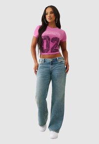 Różowy krótki t-shirt z nadrukiem graficznym i numerem "02", zestawiony z jasnoniebieskimi szerokimi jeansami i białymi trampkami.