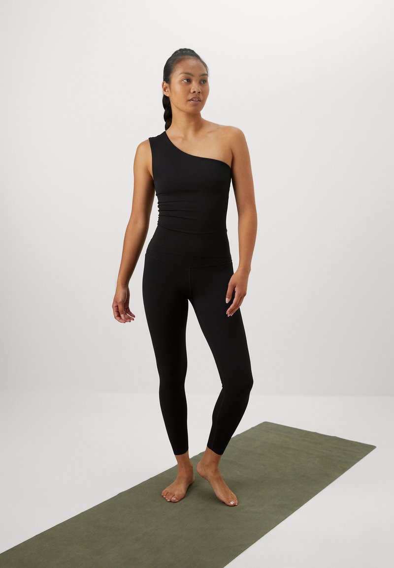 Schwarzes Top mit einem Schulterträger und taillierter Passform, kombiniert mit passenden schwarzen Leggings. Das Model steht barfuß auf einer grünen Yogamatte.