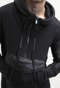 Sudadera negra con capucha alta, doble cremallera, detalles en cuero y mangas de textura acanalada. Tejido suave con un diseño estructurado.