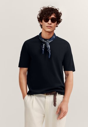 Mężczyzna o kręconych włosach, noszący czarne okulary przeciwsłoneczne, granatową apaszkę w wzory, czarną koszulę z krótkim rękawem, białe spodnie oraz brązowy pleciony pasek.