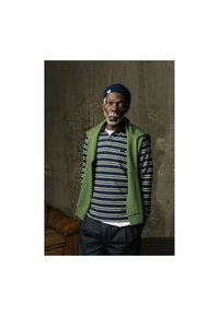 Gilet verde senza maniche sopra una polo a righe navy e bianche, abbinata a pantaloni scuri. Lo sfondo è strutturato e neutro.