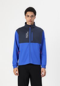 Modra in mornarsko modra zip-up jakna z visokim ovratnikom, ki ima žep na prsih, rdeče poudarke in natisnjeno logotip. Narejena iz mehkega, elastičnega materiala.