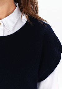 Granatowy sweter z dzianiny z szerokim dekoltem, nałożony na białą koszulę z kołnierzykiem, z dodatkowymi złotymi guzikami przy kołnierzu.