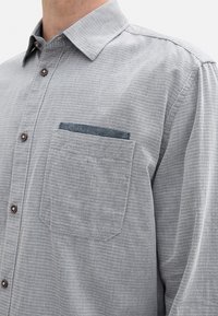 Chemise gris à manches longues avec un motif texturé, poche avant avec un accent sombre et un col standard avec des boutons bruns.