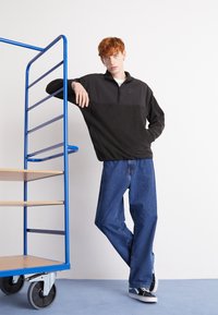 Svart fleece genser med glidelås-krage og lysegrå detaljer, kombinert med løse blå jeans og svarte joggesko, lent mot en blå vogn.