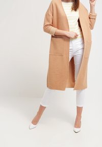 Langer, offener Cardigan aus weicher beigefarbener Wolle, mit zwei vorderen Taschen. Kombiniert mit einem gestreiften gelb-weißen Oberteil und weißen Hosen.