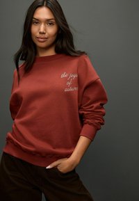 Bordový mikinový sveter s rebrovanými manžetami a spodným lemom, s bielym vyšívaným textom "radosti jesene" na ľavej hrudi. Model pózuje neformálne.