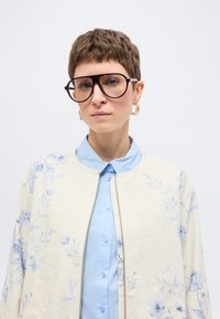 Άτομο με κοντά καστανά μαλλιά που φοράει μεγάλα στρογγυλά γυαλιά, χρυσούς κρίκους στα αυτιά, γαλάζιο πουκάμισο με κουμπιά και μπεζ σακάκι με λουλουδάτο σχέδιο.