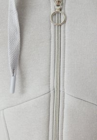 Felpa grigia chiara con cerniera, realizzata in tessuto morbido, caratterizzata da una chiusura con cerniera color argento e stringhe testurizzate sul cappuccio.