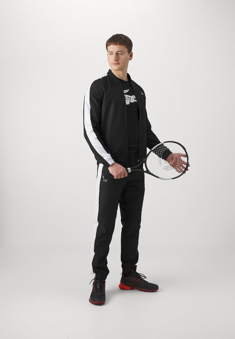 Giacca sportiva nera con strisce bianche, t-shirt nera, pantaloni neri con strisce laterali e scarpe da tennis nere/rosse, mentre tiene in mano una racchetta da tennis.