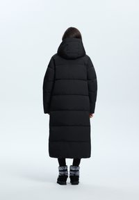 Manteau matelassé noir avec capuche, long, au design matelassé, avec des manches larges et des poignets élastiques. Porté sur des leggings noirs et des bottes à fausse fourrure.