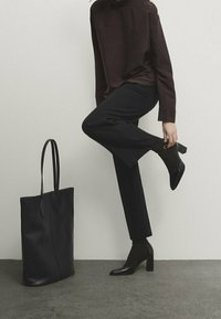 Scarpe nere con tacco, pantaloni scuri e una maglia bordeaux. Una grande borsa nera è accanto alla figura, in piedi su una superficie grigia e texture.