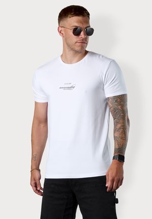 Weißes Baumwoll-T-Shirt mit Rundhalsausschnitt, versehen mit dezent bedrucktem Text auf der Brust. Das Modell trägt schwarze Sonnenbrillen und dunkle Hosen.