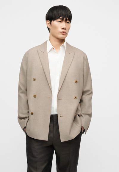 Μπεζ διπλής κοπής σακάκι με δύο καφέ κουμπιά, συνδυασμένο με λευκό πουκάμισο και σκούρο παντελόνι με μοτίβο. Λείο ύφασμα και άνετη εφαρμογή.