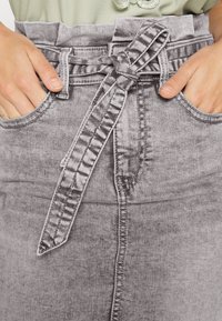 Grijze denimrok met een hoge taille, voorzien van een stoffen riem die in een strik is geknoopt, stiksel-details en twee voorzakken. De textuur lijkt zacht te zijn.