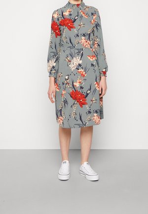 Robe fleurie avec un col montant, des manches longues et une coupe loose, ornée de fleurs rouges et blanches sur un fond gris. Portée avec des baskets blanches.