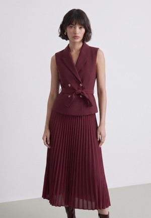 Donna che indossa blazer bordeaux senza maniche con bottoni dorati e cintura annodata, abbinato a una gonna midi plissettata coordinata.