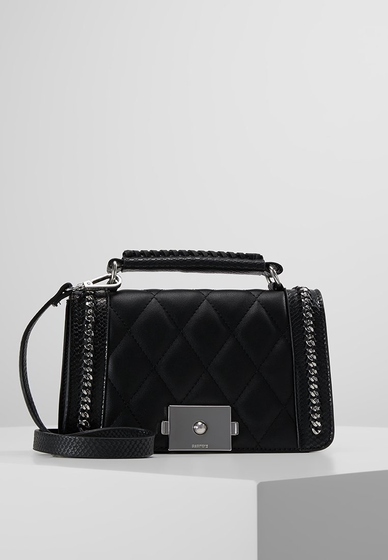 Sac à main matelassé noir avec détails en chaîne argentée, poignée supérieure et fermeture carrée argentée, placé sur un piédestal géométrique blanc sur fond gris.