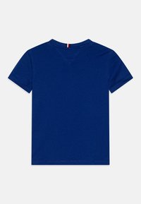 Modrá tričko s krátkým rukávem a klasickým kulatým výstřihem, hladká bavlněná textura a malý červený, bílý a modrý akcent na zadním švu výstřihu.